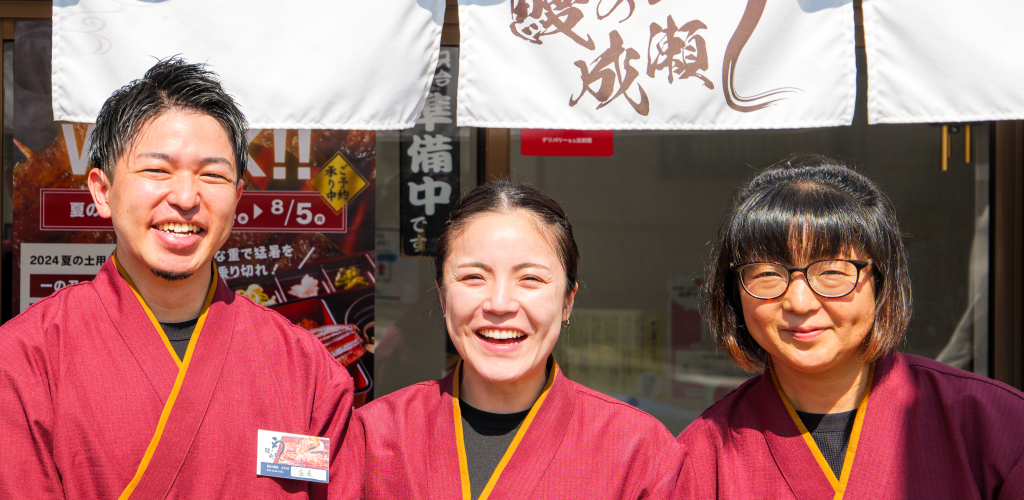 赤い作務衣を着た3人の店員が、笑顔で並んでいる様子。背後には、鰻の成瀬の暖簾が掛かっている。